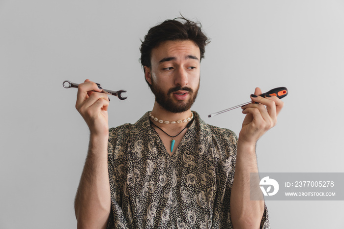 Young queer latin gay man holding a pair of tools with a doubtful expression.