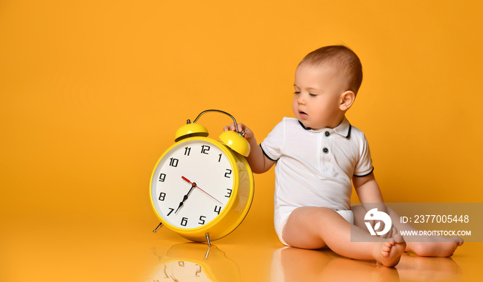 Baby boy toddler is occupied with sitting at big yellow alarm clock with hands playing showing on yellow with copy space