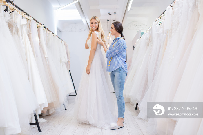 Female shop assistant helping young woman to fasten wedding dress in modern bridal store