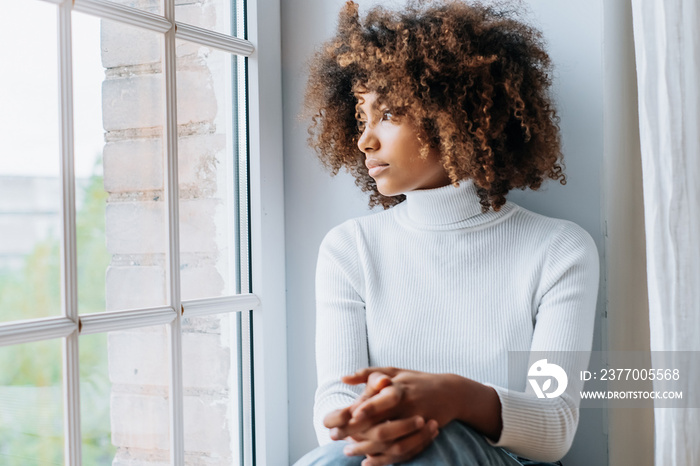 Thoughtful young woman with dark skin and modern kinky hair sits on white windowsill looking outside window close view