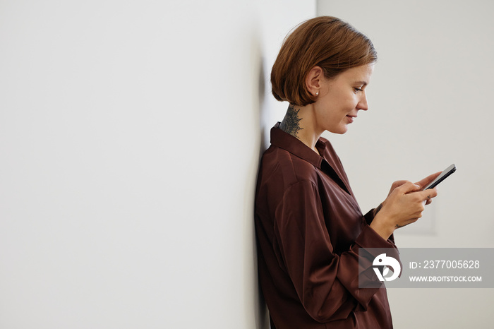 Side view waist up portrait of young tattooed woman holding smartphone while standing by white wall minimal, copy space