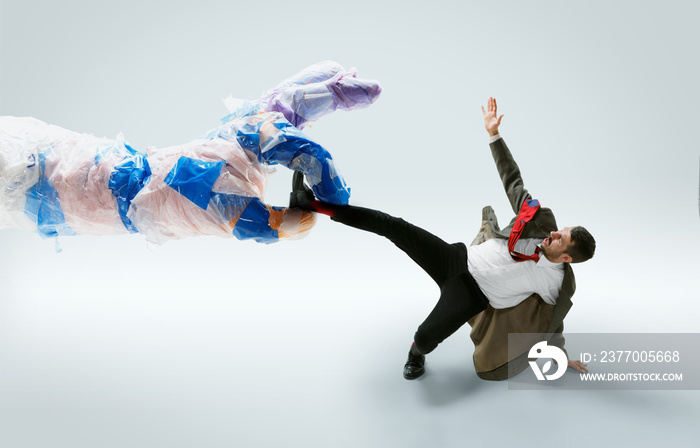 Young caucasian man avoiding big plastic hand on white studio background. Male model in office attire fighting in motion and action. Looks angry, scared, pressured. Eco problems, pollution, recycling.