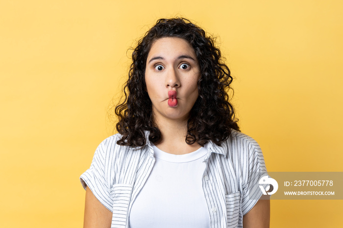 Portrait of surprised funny woman with dark wavy hair making fish face grimace with pout lips and looking with confused comical expression. Indoor studio shot isolated on yellow background.