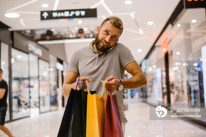 A guy with a beard walks through the mall. He carries colorful bags and talks on his cell phone