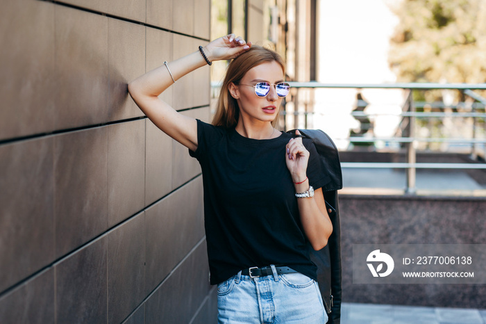 Girl wearing t-shirt, glasses and leather jacket posing against street , urban clothing style. Street photography