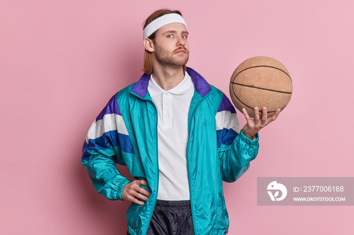 Serious man basketball player holds ball looks confidently at camera wears white headband sportsclothes enjoys playing favorite game isolated over pink background. Sport and recreation concept