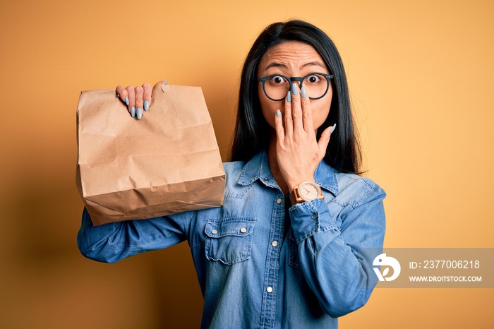 Young beautiful chinese woman holding delivery bag over isolated yellow background cover mouth with hand shocked with shame for mistake, expression of fear, scared in silence, secret concept