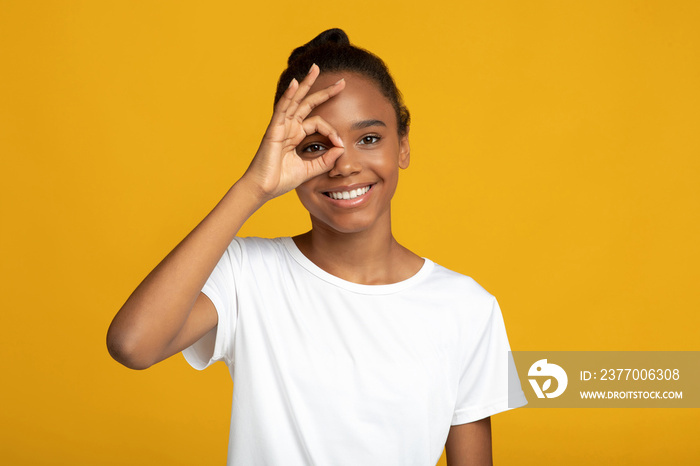 Happy teenager pretty african-american female pupil in white t-shirt makes ok gesture with hand on eye
