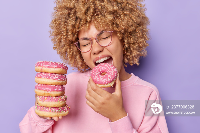 Photo of hungry sweet tooth woman with curly hair bites delicious appetizing glazed doughnut keeps mouth widely opened enjoys favorite food or snack dressed casually isolated over purple background.
