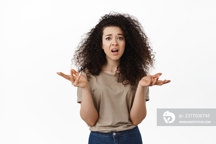 Whats going on. Confused curly woman shrugging shoudlers and raising hands puzzled, dont understand nothing, standing clueless against white background