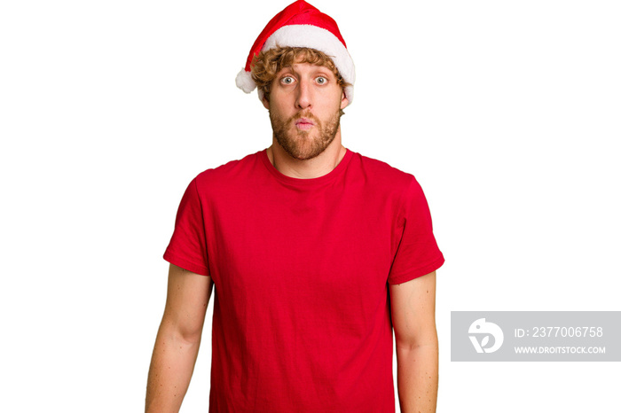 Young caucasian man wearing a Christmas Santa hat isolated on white background shrugs shoulders and open eyes confused.