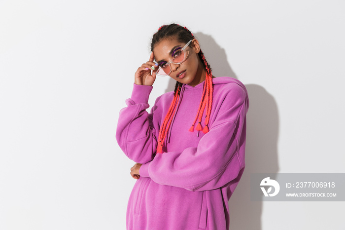 Young african stylish woman in pink sweatshirt and sunglasses posing isolated over white wall background.