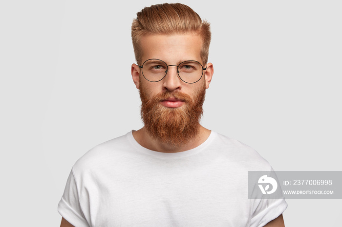 Young handsome male hipster has thick ginger beard and mustache, trendy haircut, looks seriously directly at you, wears spectacles and white t shirt, isolated over studio wall. Masculinity concept