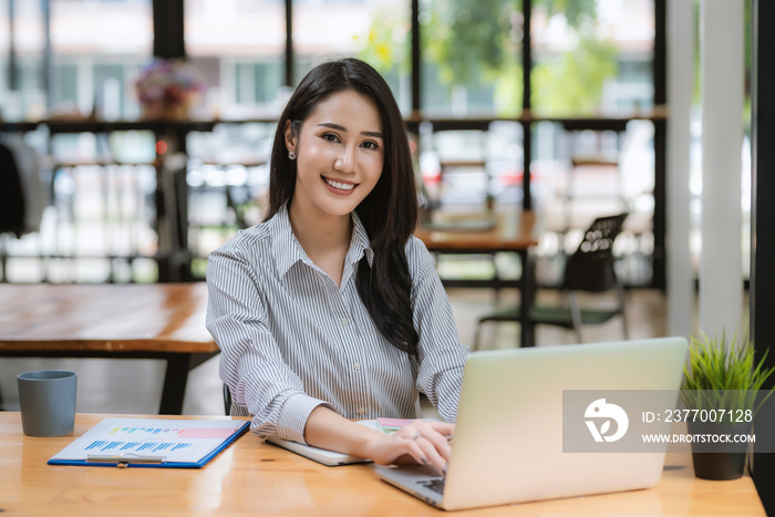 Happy young Asian businesswoman working on laptop keyboard with document at office . Looking at camera.