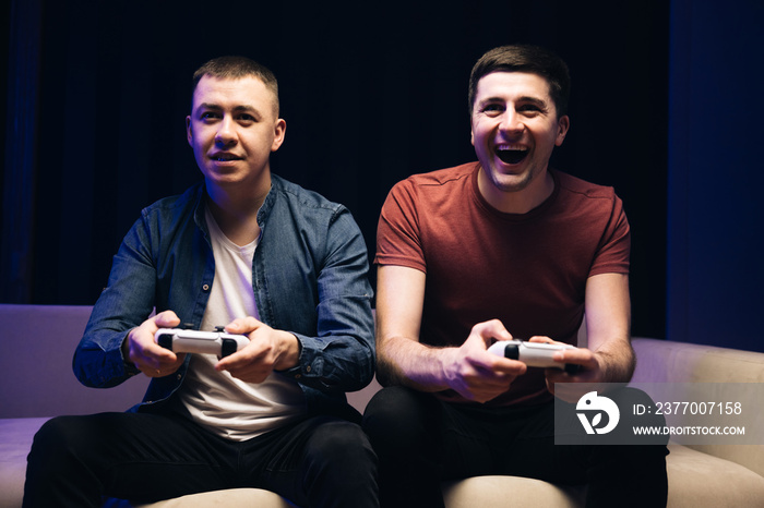 Excited funny young adult men gamers holding controllers playing video game friends winning videogame having fun together celebrating victory at home.