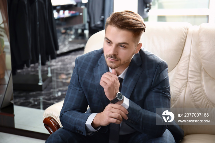 Young man in elegant suit sitting on sofa at menswear store