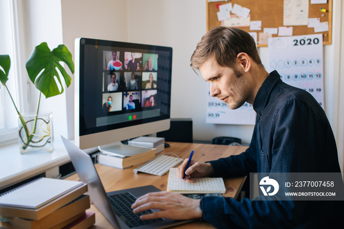 Man sitting by desktop computer and making notes. Working remotely Young man having video call via computer in the home office. Stay at home and work from home concept. Managing business team meeting