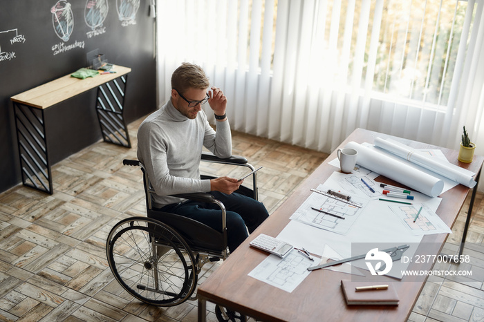 Working place. Concentrated male engineer or architect in a wheelchair working with digital tablet at his workplace in modern office