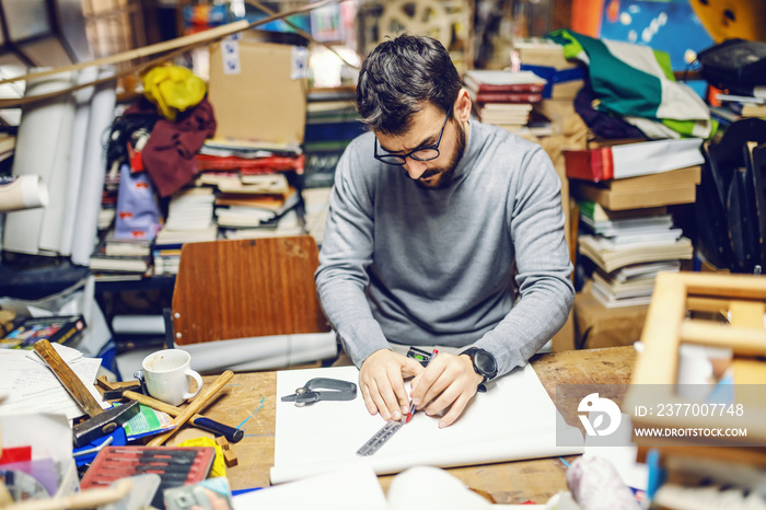 Young focused handsome Caucasian bearded bookbinder sitting in back of bookstore, using ruler and making book cover