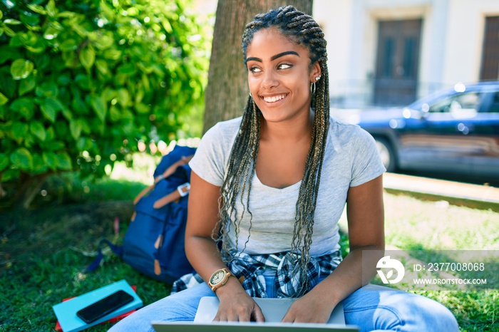 Young african american student woman smiling happy using computer laptop sitting on the grass at the university campus