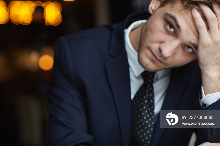 Portrait of young tired businessman wearing formal suit looking at camera with serious neutral expression resting head on hand