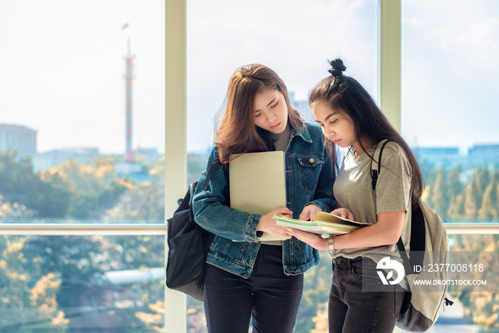 Two Asian female students discussing and pointing finger to notes in book for finding the correct answer for their question preparing for university examination. Life of studying and friendship.