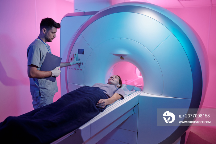 Young doctor in uniform pressing start button of mri scan machine with patient lying on long table