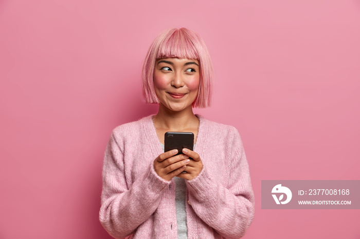 Technology, communication and lifestyle concept. Pleased dreamy woman with dyed rosy hair, holds mobile phone, uses modern app to message with friends, isolated on pink background, uses new gadget