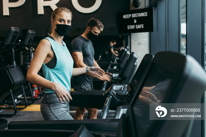 Young fit woman and man running on treadmill in modern fitness gym. They keeping distance and wearing protective face masks. Coronavirus world pandemic and sport theme.