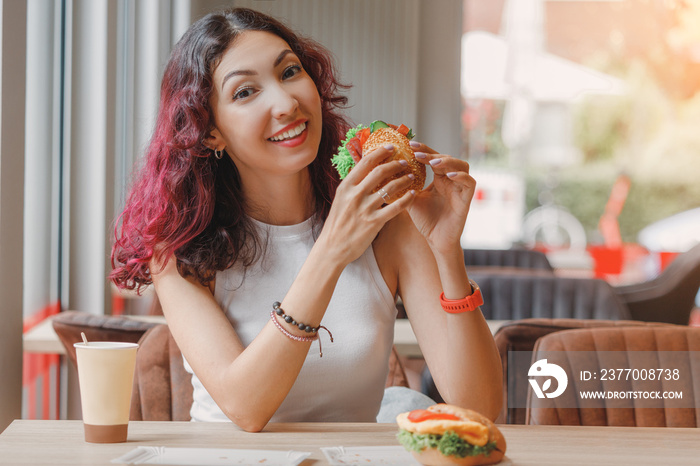 Happy girl of Asian and mixed appearance eats a hamburger or sandwich with an appetite in a fast food cafe.