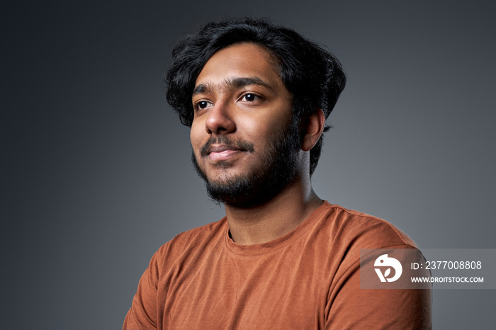 Closeup headshot of a indian guy with modern hairstyle in orange shirt he looks away and poses in gray background.