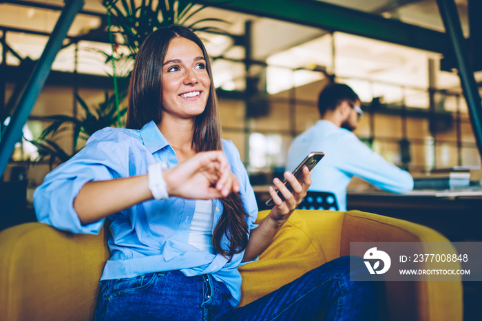 Positive successful hipster girl checking notification and gps tracking on electronic smartwatch, happy successful woman holding modern smartphone device in hand while looking away and smiling