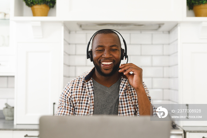 Laughing african american man sitting in kitchen making video call using laptop and headset