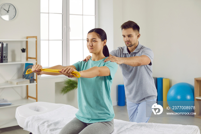 Male physiotherapist teaching female patient to do muscle rehabilitation exercises with elastic band. Young girl sitting on bed in modern physical therapy center and doing exercises with rubber band