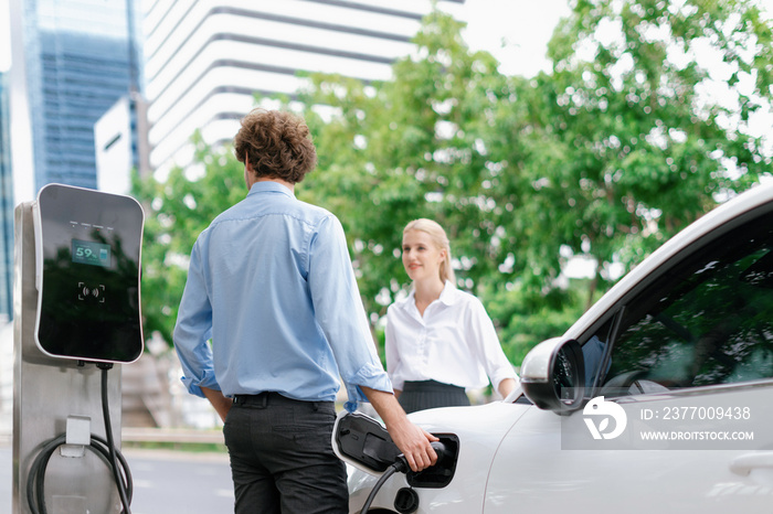 Progressive businessman and businesswoman with electric car parking and connected to public charging station before driving around city center. Eco friendly rechargeable car powered by clean energy.