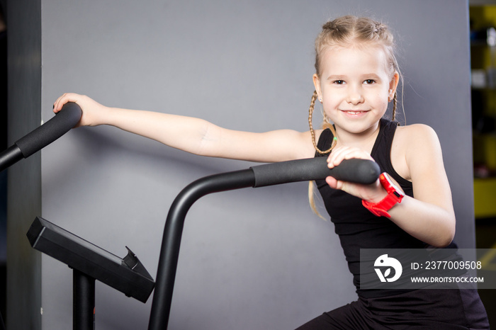 Small attractive caucasian child using exercise bike in the gym. Fitness. A little athlete using an air bike for a cardio workout at the crossfit gym. Sport girl sitting on bicycle machine