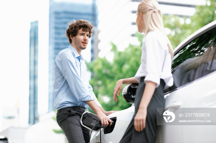 Progressive businessman and businesswoman with electric car connected to charging station before driving around city center. Eco friendly rechargeable car powered by alternative clean energy.