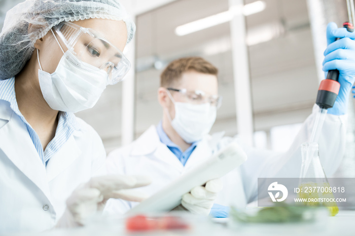 Portrait of two modern scientists wearing masks working on food research studying liquids in beaker while sitting at table in laboratory