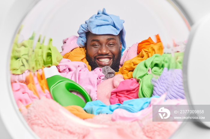 Cheerful dark skinned man covered with pile of laundry has fun poses from inside of washing machine uses detergent does househod chores at home. Washing and domestic responsibilities concept