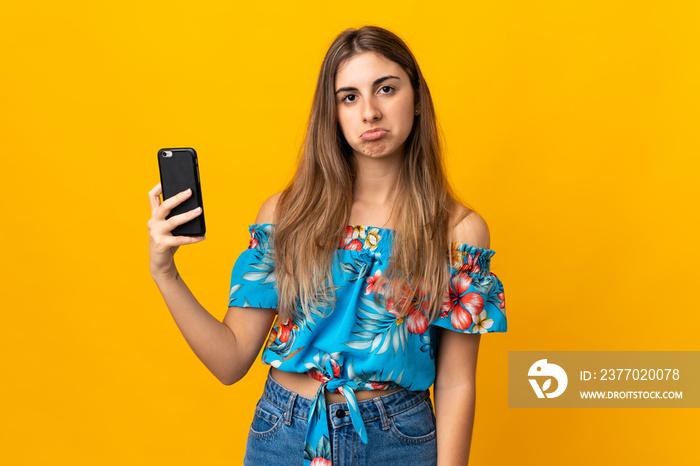 Young woman using mobile phone over isolated yellow background with sad expression