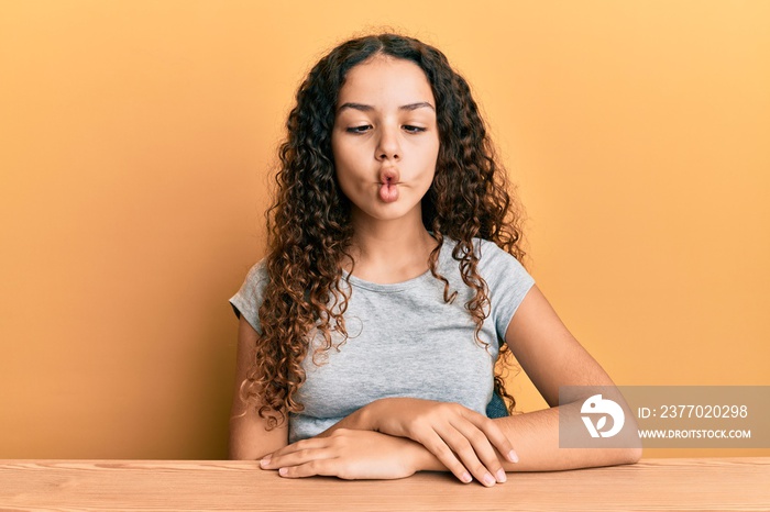 Teenager hispanic girl wearing casual clothes sitting on the table making fish face with lips, crazy and comical gesture. funny expression.