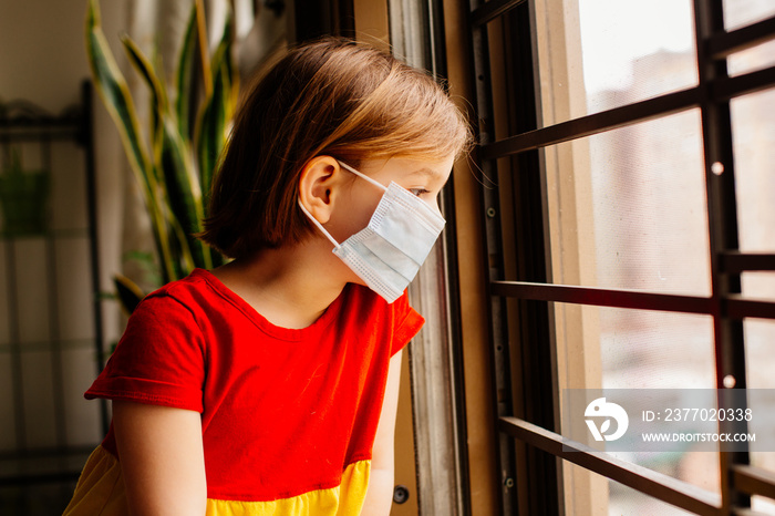 Small child with virus protection surgical face mask looking out the window, staying home for social distancing due to coronavirus