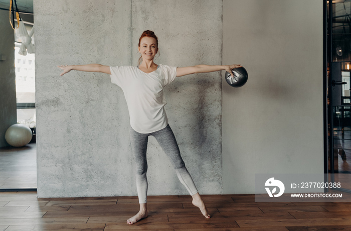 Slender beautiful red haired woman performing barre exercises in fitness studio