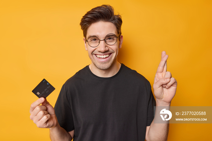 Photo of cheerful adult man keeps finger crossed believes in good luck holds credit card makes wish to recieve lump sum of money on his bank account wears round spectacles casual black t shirt