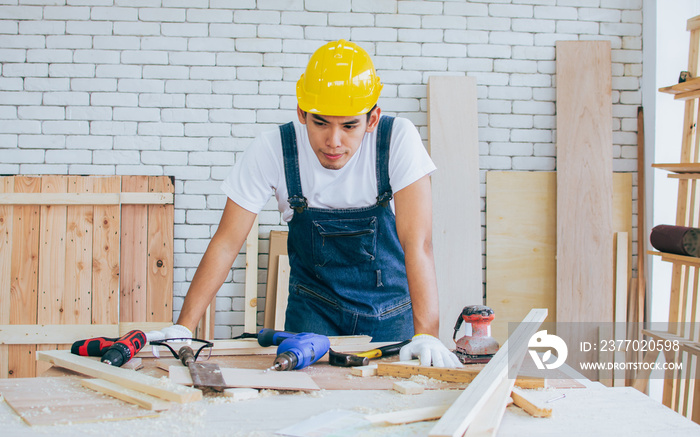 Young Asian male carpenter wearing yellow safety helmet, planning, preparing to make DIY wooden furnitures for interior decorated home. Lifestyle and Business Concept.