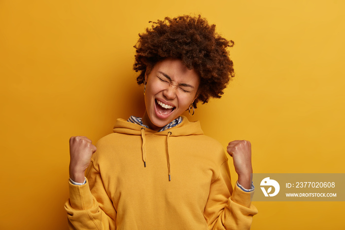 Overjoyed dark skinned woman celebrates achievement, clenches fists and feels triumph, feels empowered, watches TV game, happy favorite team scored goal, exclaims hooray, tilts head, wears hoodie