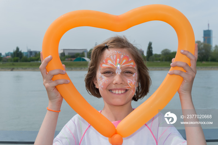 A pretty girl has a face painting and holding a balloon in shape heart