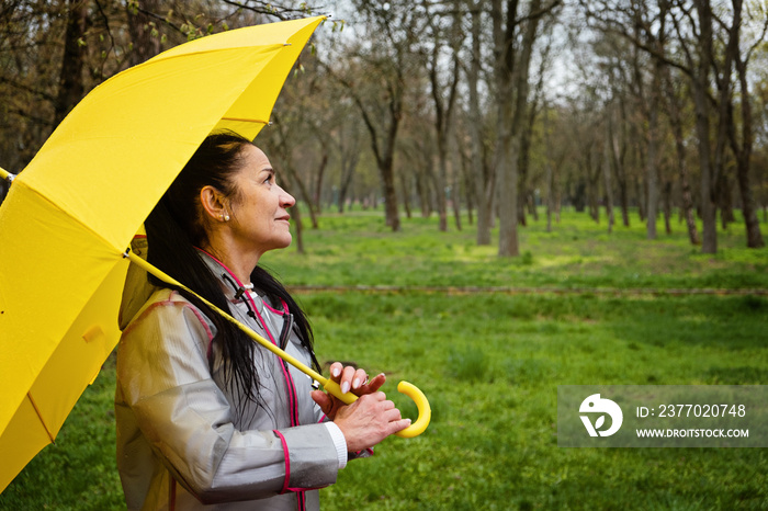 Stress resilience and mental health, concept. Managing stress and building resilience. Happy senior woman in yellow rain coat with yellow umbrella walking in park.