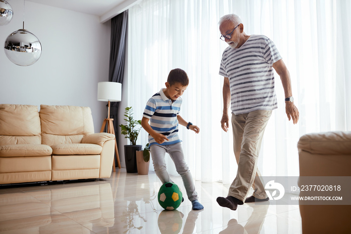 Playful little boy having fun while playing football with grandfather at home.