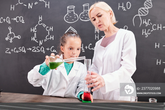 Blond confident teacher of chemistry looking at schoolgirl in whitecoat